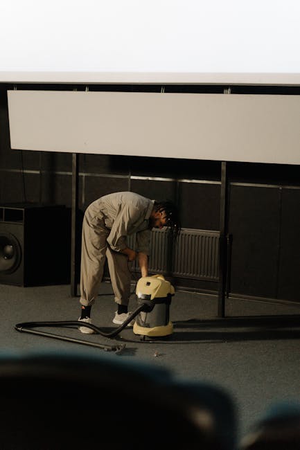 A person in a beige jumpsuit is using a vacuum cleaner for surface cleaning in a commercial or conference room, indicated by the black wall panels, radiators, and elevated white panel above. The indiv