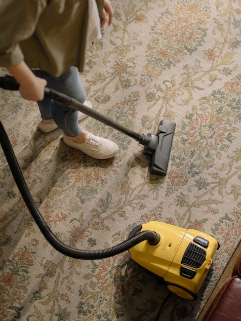 A person, wearing beige trousers and white sneakers, is using a vacuum cleaner to perform deep cleaning on an ornate, patterned carpet in a room. The carpet features a faded floral design in shades of beige, green, and orange. The vacuum cleaner is yellow with a black hose attachment, and the person is holding the metal wand with a wide cleaning head. The room has warm lighting, and the carpet appears clean with no visible stains or dirt. The scene suggests routine surface cleaning and maintenance, with Carpet Cleaning Kensington providing professional services to ensure hygienic and well-maintained carpets in residential settings.