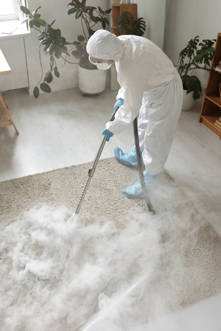 A person, wearing beige trousers and white sneakers, is using a vacuum cleaner to perform deep cleaning on an ornate, patterned carpet in a room. The carpet features a faded floral design in shades of beige, green, and orange. The vacuum cleaner is yellow with a black hose attachment, and the person is holding the metal wand with a wide cleaning head. The room has warm lighting, and the carpet appears clean with no visible stains or dirt. The scene suggests routine surface cleaning and maintenance, with Carpet Cleaning Kensington providing professional services to ensure hygienic and well-maintained carpets in residential settings.