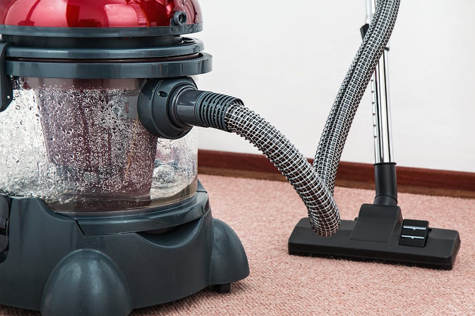 A close-up view of a commercial vacuum cleaner with a transparent dust container filled with debris, positioned on a light pink carpeted surface in a room with white walls. The vacuum hose, wrapped in a black and white patterned sleeve, extends to a floor attachment resting on the carpet. The room features a wooden baseboard along the bottom of the wall, and bright lighting highlights the cleanliness of the carpet and the equipment. The image emphasizes surface cleaning and maintenance as part of professional carpet deep cleaning services offered by Carpet Cleaning Kensington, integrating visual cues related to hygiene and cleaning tools in a domestic setting.
