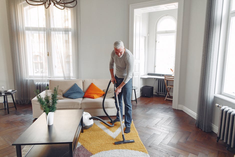 A man with gray hair and a beard is performing surface cleaning in a bright, modern living room. He is vacuuming a multicolored patterned rug with a canister vacuum cleaner. The room features large windows with semi-transparent curtains, allowing natural light to illuminate the space. There is a white sofa with blue and orange cushions, a dark-colored coffee table with a potted plant, and a wooden side table with a few items on it. The flooring consists of polished dark wooden parquet, and the walls are painted white, creating a clean and fresh atmosphere. The scene demonstrates thorough domestic cleaning practices, highlighting surface cleaning and maintenance, and is indicative of the professional services provided by Carpet Cleaning Kensington. The natural light and tidy arrangement emphasize a hygienic and well-maintained environment suitable for detailed deep cleaning and sanitisation efforts. The overall setting reflects a focus on hygiene, cleanliness, and ongoing maintenance of residential living spaces, aligned with the Kensington High Street carpet cleaning guide W8 tips.