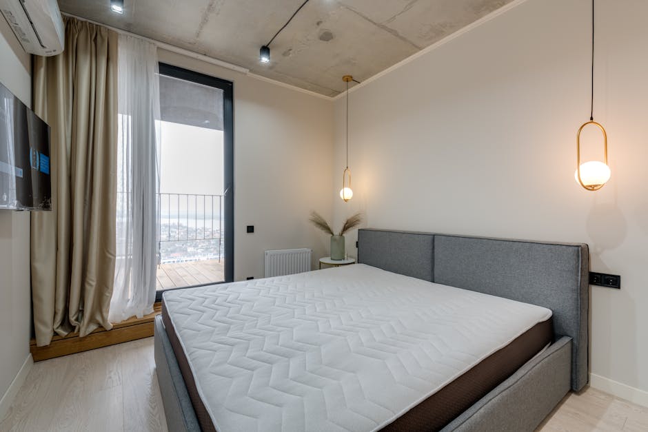 A modern bedroom featuring a neatly made bed with a light grey upholstered headboard and white mattress, positioned against a plain white wall. To the left, beige curtains are drawn open, revealing a glass door leading to a balcony with a metal railing, allowing natural light to illuminate the room. A small bedside table with a decorative vase containing dried pampas grass is situated near the bed. Two pendant lights with warm glow hang from the ceiling on either side of the bed, providing soft ambient lighting. The room has a minimalist design with light wooden flooring, and visible elements such as a wall-mounted television and a radiator add to the contemporary aesthetic. This image showcases a clean, well-maintained residential space suitable for professional domestic cleaning by Carpet Cleaning Kensington, aligned with the surface cleaning and sanitisation themes in the Notting Hill Gate mattress cleaning W11 local guide.