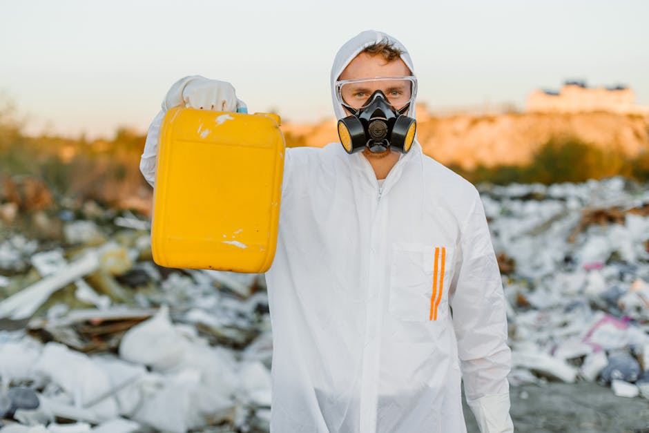 A person wearing protective gear, including a white hazmat suit, gloves, goggles, and a respirator mask, is holding a clear spray bottle filled with liquid, appearing to perform environmental testing 