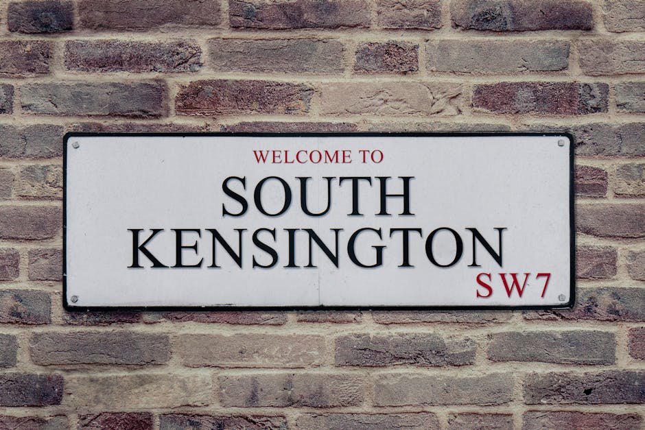 A rectangular white street sign mounted on a red brick wall, displaying the words 'WELCOME TO SOUTH KENSINGTON' in large black letters, with 'SW7' in red at the bottom right corner. The brick wall has a textured surface with a mix of light and dark reddish-brown bricks. The sign appears clean and well-maintained, with small visible screw holes at each corner for mounting, and is evenly illuminated by soft, natural lighting, emphasizing the clarity and prominence of the text. This setting suggests a typical urban environment in South Kensington, London, associated with the area’s residential and commercial properties. Carpet Cleaning Kensington offers surface and deep cleaning services that ensure a hygienic and polished appearance of such signage and surrounding surfaces.