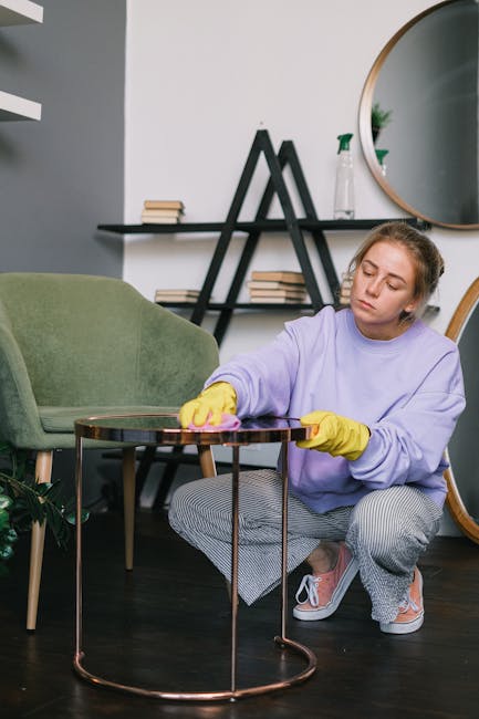 A young woman with blonde hair tied back, wearing a lavender sweatshirt, yellow cleaning gloves, and grey checkered pants, is kneeling on a dark wooden floor while thoroughly cleaning a round glass side table with a pink cloth. The room features a modern aesthetic with a light grey wall, a green upholstered armchair, a black triangular shelf with books, and a large round mirror mounted on the wall reflecting part of the space. Bright, natural lighting illuminates the scene, emphasizing the cleanliness and neatness of the area. The setting appears to be a contemporary living room, with attention to surface cleaning and maintaining hygiene, characteristic of domestic deep cleaning services provided by Carpet Cleaning Kensington, as indicated on the page about South Kensington upholstery cleaning near Victoria and Albert.